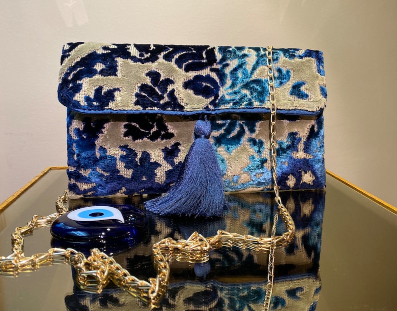 Op de afbeelding: Een blauwe en zilveren velvet clutch met een gouden kettingband en een blauwe kwast. De tas heeft een blauwe kwade oog charme.