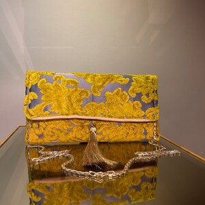 K&ouml;nnte beinhalten: Eine goldene und violette Clutch aus Samt mit einer goldenen Kettenkette und einem Quastenanh&auml;nger. Die Tasche hat ein Blumenmuster.