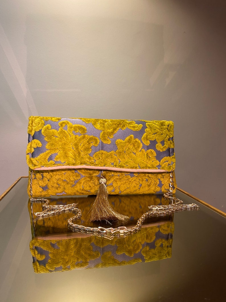 K&ouml;nnte beinhalten: Eine goldene und violette Clutch aus Samt mit einer goldenen Kettenkette und einem Quastenanh&auml;nger. Die Tasche hat ein Blumenmuster.