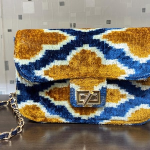 May include: A rectangular handbag with a gold chain strap. The bag features a geometric pattern in blue, gold, and white. The clasp is gold-toned. The bag is made of a soft, velvety material.