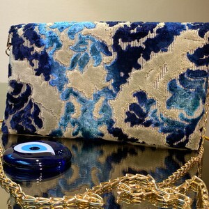 Op de afbeelding: Een blauwe clutch met een wit patroon, een gouden kettingriem en een blauwe hanger in de vorm van een oog.