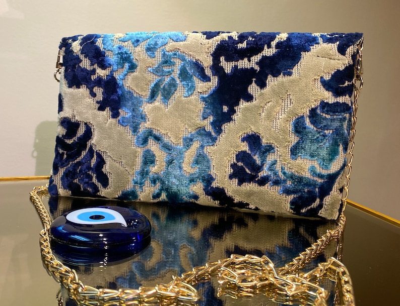 Op de afbeelding: Een blauwe clutch met een wit patroon, een gouden kettingriem en een blauwe hanger in de vorm van een oog.