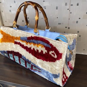 Op de afbeelding: Een rechthoekige draagtas met een bamboe handvat en een kleurrijk patroon. De tas heeft een lichtblauwe binnenkant en een textuurrijke buitenkant met rode, blauwe en gouden accenten. De handvatten zijn van gebogen bamboe.