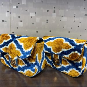 May include: Two rectangular handbags with a gold, blue, and white geometric pattern. The bags have a flap closure with a gold-colored clasp and a gold chain strap. The bags are made of a soft, velvet-like material.