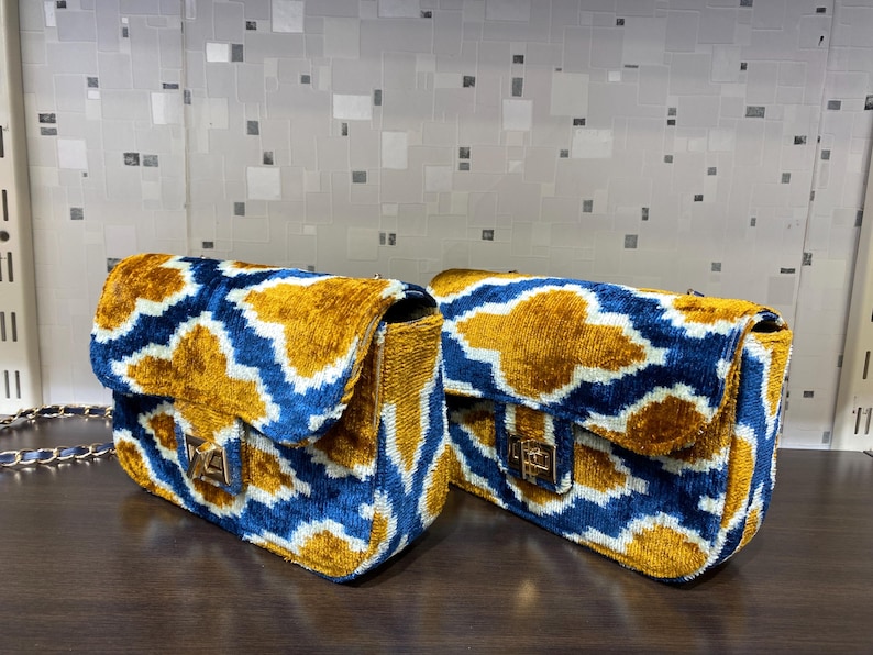 May include: Two rectangular handbags with a gold, blue, and white geometric pattern. The bags have a flap closure with a gold-colored clasp and a gold chain strap. The bags are made of a soft, velvet-like material.
