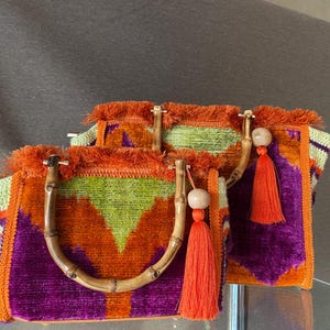 May include: Two colorful crocheted handbags with fringe details and bamboo handles. The bags are made with a mix of orange, green, purple, and cream colored yarn.