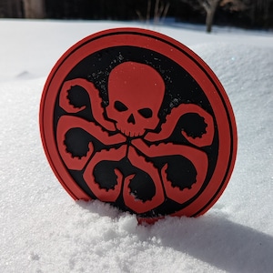 Puede incluir: Una pegatina circular roja y negra con un diseño de calavera y tentáculos. La pegatina está en la nieve.