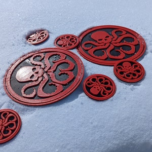 Puede incluir: Una colecci&oacute;n de pegatinas circulares rojas y negras con un dise&ntilde;o de calavera y pulpo, colocadas sobre una superficie blanca nevada.