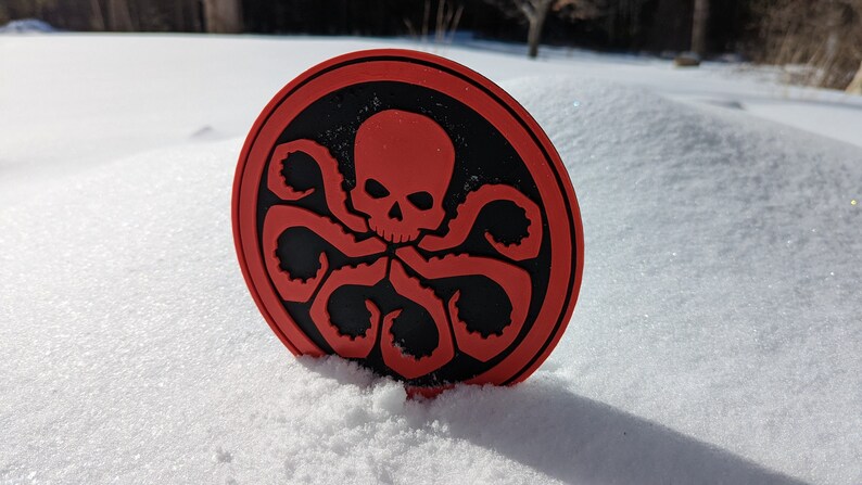 Puede incluir: Una pegatina circular roja y negra con un dise&ntilde;o de calavera y tent&aacute;culos. La pegatina est&aacute; en la nieve.