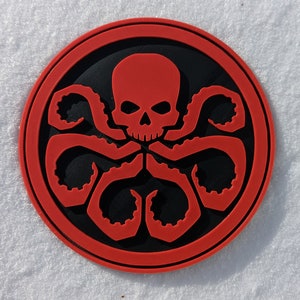 Puede incluir: Una pegatina circular roja y negra con un cr&aacute;neo y tent&aacute;culos, que se asemeja al logotipo de la organizaci&oacute;n ficticia HYDRA.