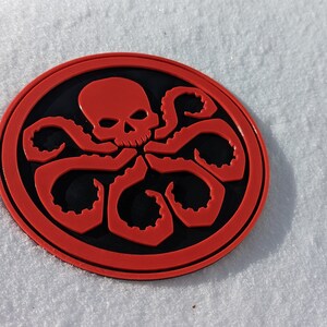 Puede incluir: Un parche circular rojo y negro con una calavera y tent&aacute;culos, que se asemeja al logotipo de una organizaci&oacute;n ficticia.