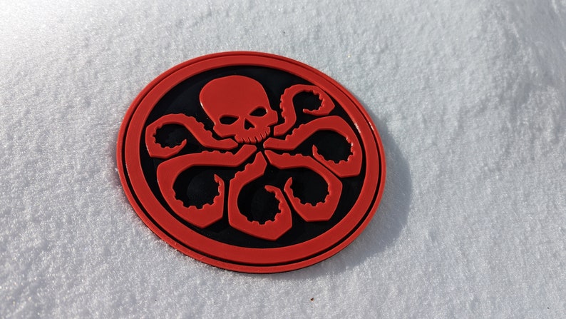 Puede incluir: Un parche circular rojo y negro con una calavera y tent&aacute;culos, que se asemeja al logotipo de una organizaci&oacute;n ficticia.