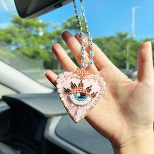 Könnte beinhalten: Ein rosa herzförmiger Autoanhänger mit einem gestickten Augendesign. Der Anhänger ist mit Perlen und Perlen verziert und hängt an einer Perlenkette. Das Auge hat blaue, weiße und goldene Akzente.