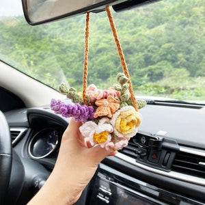 Pode incluir: Um buquê de flores de crochê pendurado no espelho retrovisor de um carro. O buquê é feito de fio rosa, branco e roxo e é um acessório de carro encantador.