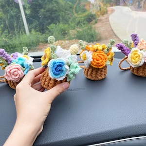 May include: Crocheted flower arrangements in small brown pots. The flowers are in various colors, including pink, blue, yellow, and orange. The pots have a small loop for hanging. The arrangement is on a gray surface.