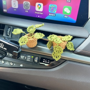Könnte beinhalten: Zwei gehäkelte grüne Pflanzendekorationen in braunen Töpfen, platziert auf dem Armaturenbrett eines Autos. Das Armaturenbrett des Autos zeigt 20,3 auf einem digitalen Display.