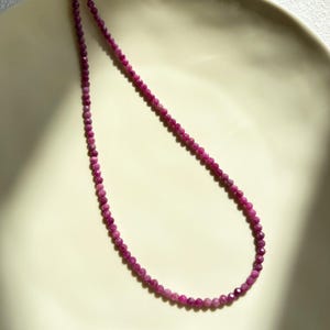 Puede incluir: Un collar de una sola hilera de pequeñas cuentas facetadas de color magenta intenso dispuestas en línea curva. Las cuentas tienen una forma circular uniforme y una superficie reflectante, creando una textura consistente a lo largo de la pieza.