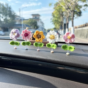 May include: Five crocheted flower decorations in different colors, including purple, pink, yellow, white, and pink, are displayed on a car dashboard. The flowers are attached to small green stems.
