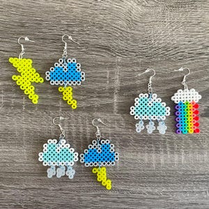 May include: Five pairs of colorful beaded earrings with weather-themed designs. The earrings feature a lightning bolt, a cloud with raindrops, a rainbow, and two clouds with lightning bolts.