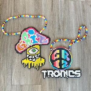 May include: Two colorful beaded necklaces with unique designs. One features a mushroom shape with a melting yellow base and an eye. The other is a circle with the letters "GB" and "12". The word "TRONICS" is below the circle.