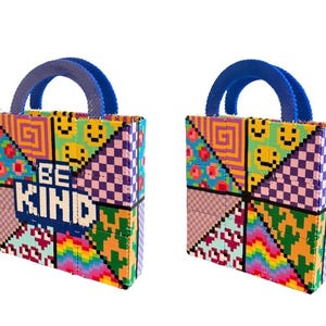 May include: Two colorful tote bags with blue handles. The bags feature a pixelated design with various patterns, including smiley faces, flowers, and geometric shapes. The words "BE KIND" are prominently displayed on the front of the bag.