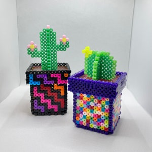 May include: Two small potted cactus plants made of colorful plastic beads. The cactus on the left is green with pink flowers and is in a black pot with a colorful geometric pattern. The cactus on the right is green with a yellow flower and is in a purple pot with a colorful mosaic pattern.