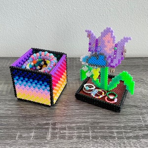 May include: A rainbow-colored cube-shaped box with a flower-shaped jewelry holder. The flower is purple with green leaves and a blue center. The box and holder are made of plastic beads.