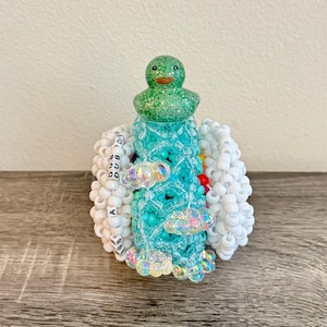May include: A whimsical beaded sculpture featuring a green glitter duck head atop a turquoise and white beaded structure. The sculpture includes iridescent cloud-shaped beads and white beads with the letters "DUB" and "A".