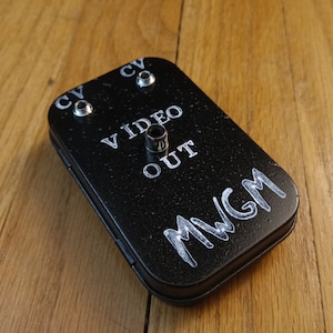 Puede incluir: Una caja de metal negra con las palabras "VIDEO OUT" y "MWGM" escritas en ella. Hay dos pequeños botones plateados etiquetados como "CV" en la parte superior de la caja.