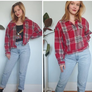 May include: A woman is wearing a red and black checked flannel shirt with a cropped cut. She is also wearing light blue jeans and black boots.