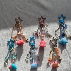 May include: A collection of handmade keychains with star-shaped metal toppers. Each keychain features a unique charm, including pink octopus, blue sea creatures, and other colorful beads. The keychains are displayed on a white, textured surface.