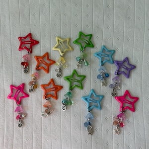 May include: A selection of star-shaped hair clips in a range of colours: red, orange, yellow, green, blue, and pink. Each clip features a dangling charm with beads and star details. The clips are displayed on a white, textured background.