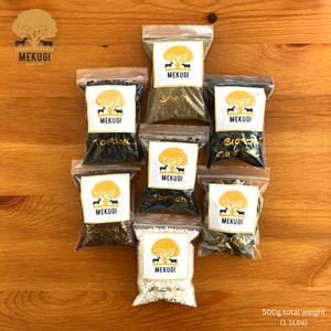 Pode incluir: Oito pequenos sacos de plástico transparente cheios de diferentes tipos de terra. Cada saco tem um rótulo branco com um logotipo dourado e o texto "MEKUGI". Os sacos estão dispostos em uma grade sobre uma superfície de madeira. O texto "500g total weight (1.1Lbs)" é impresso abaixo dos sacos.