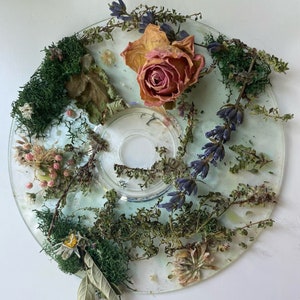 Może przedstawiać: Przezroczysty dysk z różnymi suszonymi kwiatami i zielenią ułożonymi na górze. Kwiaty obejmują różową różę, lawendę i małe białe kwiaty. Zieleń obejmuje mech i różne rodzaje liści.