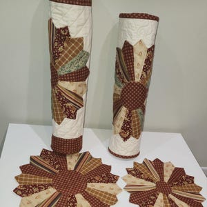May include: Set of three quilted table runners and placemats in a brown, beige, and green patchwork pattern. The runners are rolled up and the placemats are laid flat. The pattern features a large flower design in the center.
