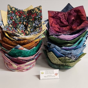 May include: A stack of colorful fabric bowl cozies in various patterns, including floral, abstract, and geometric designs. The cozies are designed to hold bowls of hot food and keep hands safe from heat. A small card with instructions is also visible.