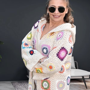 Puede incluir: Una chaqueta de punto de ganchillo de color crema con capucha, que presenta coloridos patrones circulares en tonos de rosa, morado, azul, verde, amarillo y naranja.