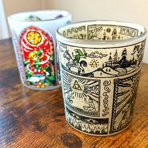 May include: Two glass candle holders with a Zelda theme. The holder on the left has a red, green, and yellow design with a character from the game. The holder on the right has a black and white design with a character riding a horse.