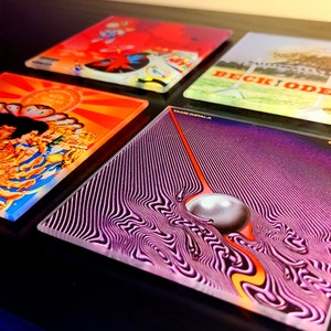 May include: Four different vinyl record coasters. The coasters are square and feature different album covers. The top left coaster is red with a cartoon character. The top right coaster is green with a tree and the text "Beck, Ode." The bottom left coaster is orange with a colourful illustration. The bottom right coaster is purple with a swirling pattern and a white orb.