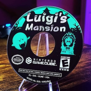 May include: A green and black Nintendo GameCube game disc with the title "Luigi's Mansion" printed on it. The disc is in a clear plastic case with a stand. The ESRB rating of "Everyone" is visible on the disc.