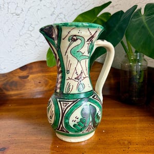 May include: A hand-painted ceramic pitcher with a cream-colored base, green and brown accents, and a green bird design. The pitcher has a handle and a flared rim. The design is reminiscent of folk art.