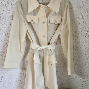 May include: Cream-colored trench coat with a matching belt. The coat features a collared neckline, button-down front, and two front pockets. The coat is hanging on a black hanger against a textured white wall.