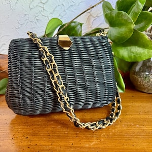 May include: Black woven handbag with a gold chain strap and clasp. The rectangular purse has a textured, basket-weave design. The gold chain is intertwined with black leather. The bag is set against a wooden surface with green foliage in the background.
