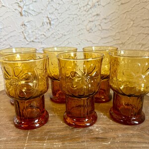 May include: Set of amber-colored drinking glasses. Each glass features a floral design on the bowl and a pedestal base. The glasses are arranged on a wooden surface, showcasing their vintage aesthetic.