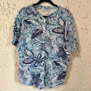 May include: A short-sleeved button-up shirt with a floral pattern in shades of blue and white. The shirt features a crew neck and white buttons down the front. The floral design includes large flowers and leaves.