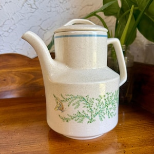 May include: Cream-colored ceramic teapot with a lid and handle. The teapot features a blue stripe around the lid and a green and yellow butterfly and plant design. The teapot is sitting on a wooden surface.
