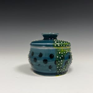 Op de afbeelding: Een handgemaakte keramische pot met een deksel. De pot is teal met zwarte stippen en witte getextureerde accenten. Het deksel is een donkerdere teal met witte getextureerde details. De pot is een decoratieve opbergcontainer.