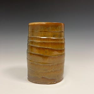 May include: A handcrafted ceramic tumbler with a textured, ridged design. The cylindrical cup is glazed in shades of brown and gold, creating a warm, earthy aesthetic. The pottery piece is suitable for beverages and decorative use.