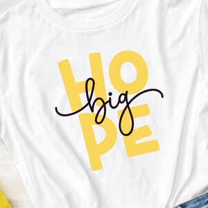 May include: White t-shirt with the words "HOPE big" printed in yellow and black. The shirt is laying flat on a wooden surface.