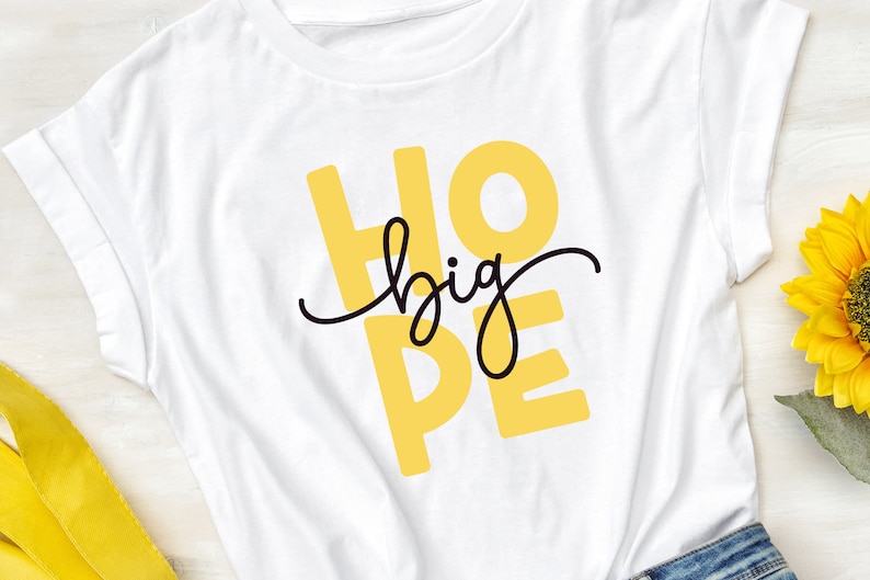 May include: White t-shirt with the words "HOPE big" printed in yellow and black. The shirt is laying flat on a wooden surface.